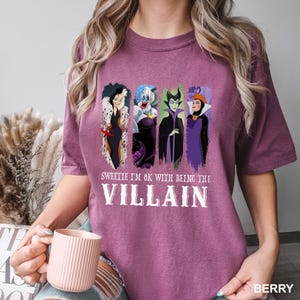 Puede incluir: Camiseta color bayas con cuatro personajes de villanos de dibujos animados. La camiseta tiene el texto "Sweetie I'm OK with being the Villain" en letras blancas. La camiseta es de cuello redondo y manga corta.