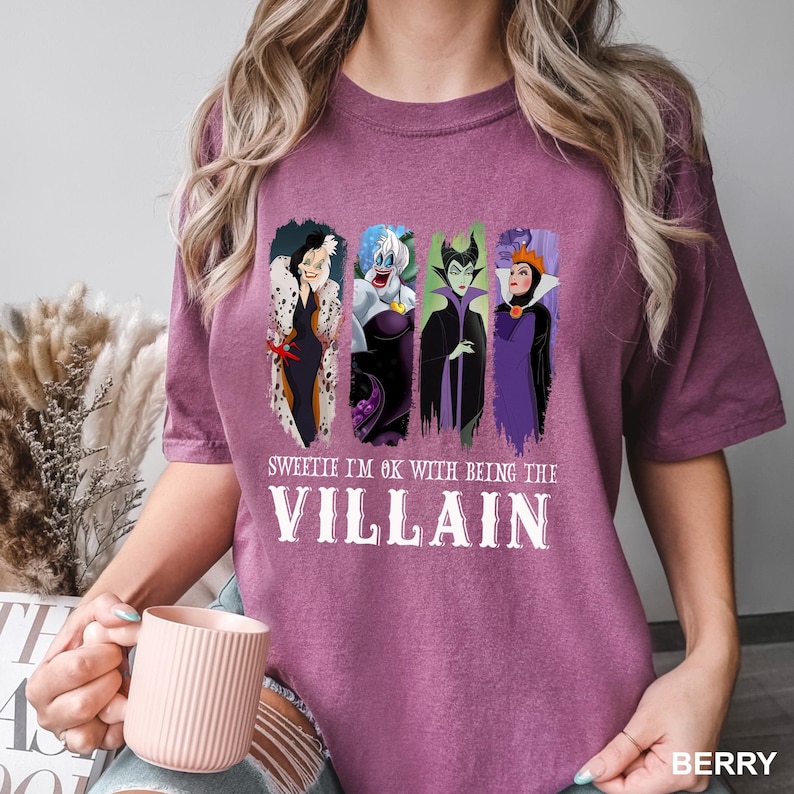 Puede incluir: Camiseta color bayas con cuatro personajes de villanos de dibujos animados. La camiseta tiene el texto "Sweetie I'm OK with being the Villain" en letras blancas. La camiseta es de cuello redondo y manga corta.