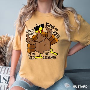 May include: Mustard-colored t-shirt featuring a cartoon turkey wearing sunglasses. The turkey has its wings up and the text reads "Turkey Gravy, Beans and Rolls, Let Me See That Casserole". A fun, festive design for Thanksgiving.
