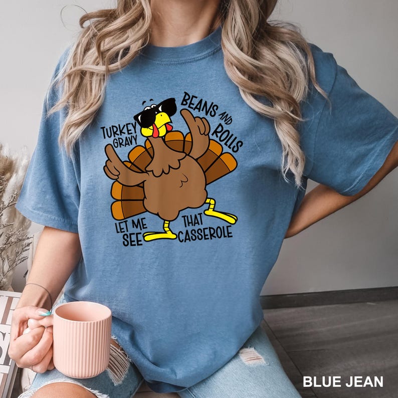 May include: Blue jean colored t-shirt featuring a cartoon turkey wearing sunglasses and pointing. The shirt has the phrases "Turkey Gravy", "Beans and Rolls", "Let Me See", and "That Casserole" printed on it. The shirt is a comfortable, casual style.