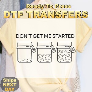 May include: A light yellow t-shirt with the text "DON'T GET ME STARTED" above a graphic of three jars. The jars have varying amounts of contents. The words "Ready To Press DTF TRANSFERS" are at the top.