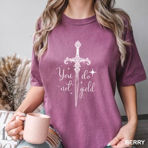 Peut inclure: T-shirt de couleur baie avec un motif blanc d'&eacute;p&eacute;e et le texte "You do not yield". Le t-shirt a des manches courtes et une coupe d&eacute;contract&eacute;e. L'image montre &eacute;galement une personne tenant une tasse rose.