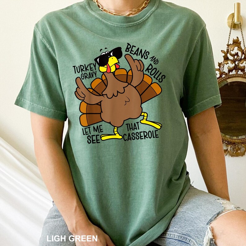 May include: Light green t-shirt featuring a cartoon turkey wearing sunglasses and gesturing with its wings. The text on the shirt reads "Turkey Gravy, Beans and Rolls, Let Me See That Casserole."