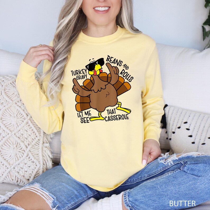 May include: A light yellow long-sleeve shirt with a cartoon turkey graphic wearing sunglasses. The turkey has brown feathers and is giving the middle finger. Text on the shirt says "Turkey Gravy, Beans and Rolls, Let Me See That Casserole."