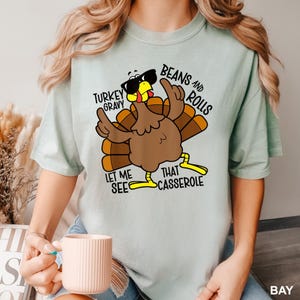May include: A light green t-shirt featuring a cartoon turkey wearing sunglasses and pointing upwards. The text on the shirt reads "Turkey Gravy, Beans and Rolls, Let Me See That Casserole."