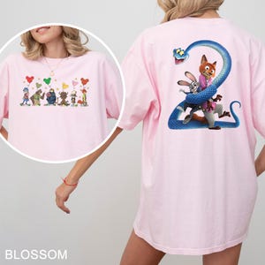 K&ouml;nnte beinhalten: Hellrosa T-Shirt mit Cartoon-Illustrationen auf Vorder- und R&uuml;ckseite. Die Vorderseite zeigt Cartoon-Charaktere mit herzf&ouml;rmigen Luftballons. Die R&uuml;ckseite zeigt eine blaue Schlange, die sich um Cartoon-Charaktere wickelt. Das Wort "BLOSSOM" steht unten.