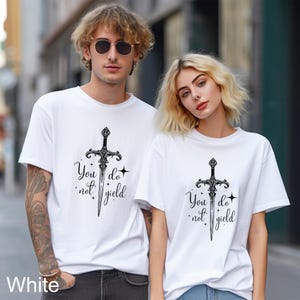 Peut inclure: T-shirts blancs avec un motif d'&eacute;p&eacute;e noire et le texte "You do not yield". Les t-shirts sont &agrave; manches courtes et ont un col rond. Le motif est centr&eacute; sur le devant.