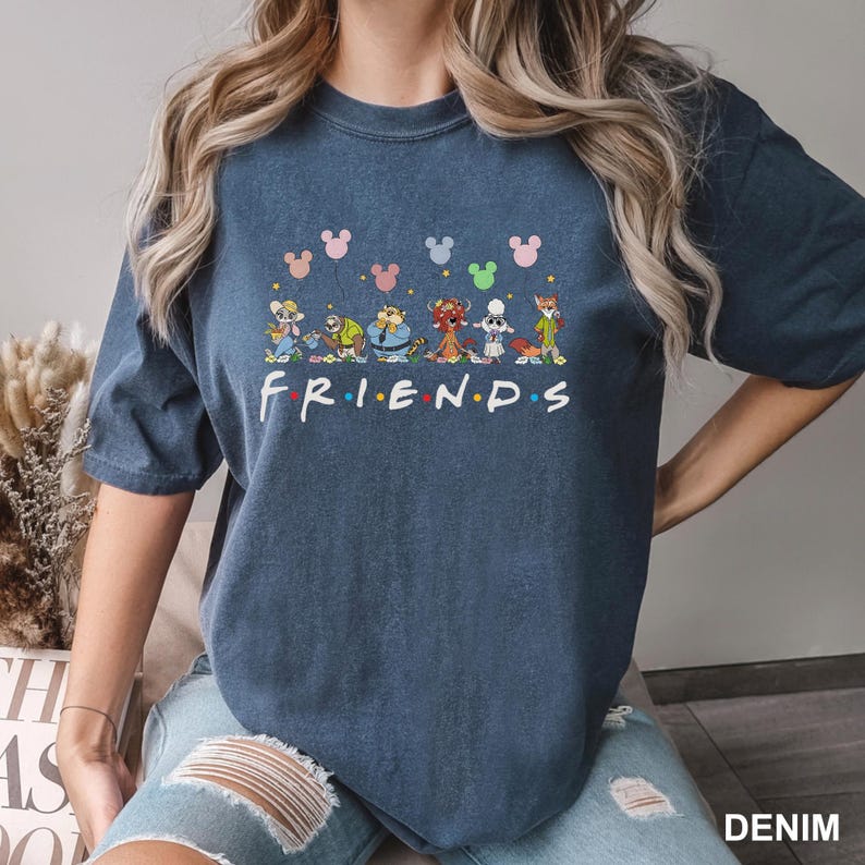 May include: A denim blue t-shirt featuring a colorful graphic of cartoon characters and the word "FRIENDS" in a playful font. Above the characters are Mickey Mouse head silhouettes in various colors. The shirt is a casual, comfortable style.