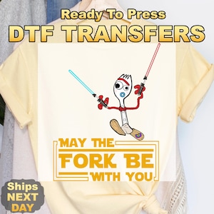 May include: Cream-colored t-shirt with a DTF transfer featuring a cartoon fork character wielding lightsabers. The text "MAY THE FORK BE WITH YOU" is below the character. The shirt is ready to press.