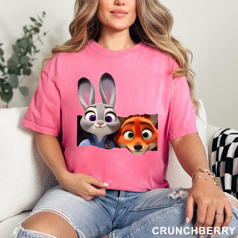 May include: Pink t-shirt featuring a graphic of Judy Hopps and Nick Wilde from Zootopia. The cartoon characters are set against a torn paper background. The word "CRUNCHBERRY" is printed in white at the bottom of the shirt.