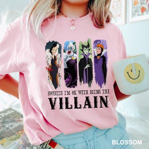 Puede incluir: Camiseta rosa claro con un estampado de cuatro villanas de Disney: Cruella de Vil, &Uacute;rsula, Mal&eacute;fica y la Reina Malvada. El texto "Sweetie I'm OK with being the Villain" est&aacute; impreso debajo. Una taza blanca con una carita sonriente amarilla est&aacute; en la imagen.