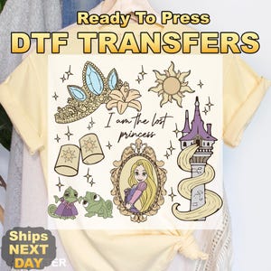 May include: A light yellow t-shirt featuring DTF TRANSFERS. The design includes a princess tiara, sun, flower, and a portrait of a princess with the text "I am the lost princess". Also includes two frogs and a tower.