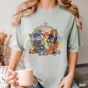 Pode incluir: Camiseta verde claro com um gr&aacute;fico colorido de personagens do filme de anima&ccedil;&atilde;o Zootopia. O design inclui Judy Hopps, Nick Wilde e outros personagens, rodeados por flores e bal&otilde;es. A palavra "BAY" est&aacute; impressa no canto inferior direito.