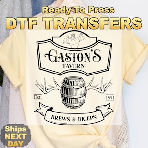 May include: A light yellow t-shirt with a black graphic design. The design includes the text "Gaston's Tavern", a barrel, antlers, and the words "Brews & Biceps". The shirt also has the text "Ready To Press DTF TRANSFERS".