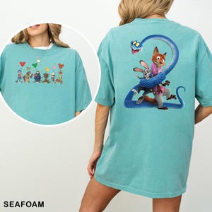 K&ouml;nnte beinhalten: T&uuml;rkisgr&uuml;nes T-Shirt mit Cartoon-Grafiken auf Vorder- und R&uuml;ckseite. Die Vorderseite zeigt Cartoon-Figuren mit herzf&ouml;rmigen Luftballons. Die R&uuml;ckseite zeigt einen Fuchs und ein Kaninchen, die von einer blauen Schlange umarmt werden.