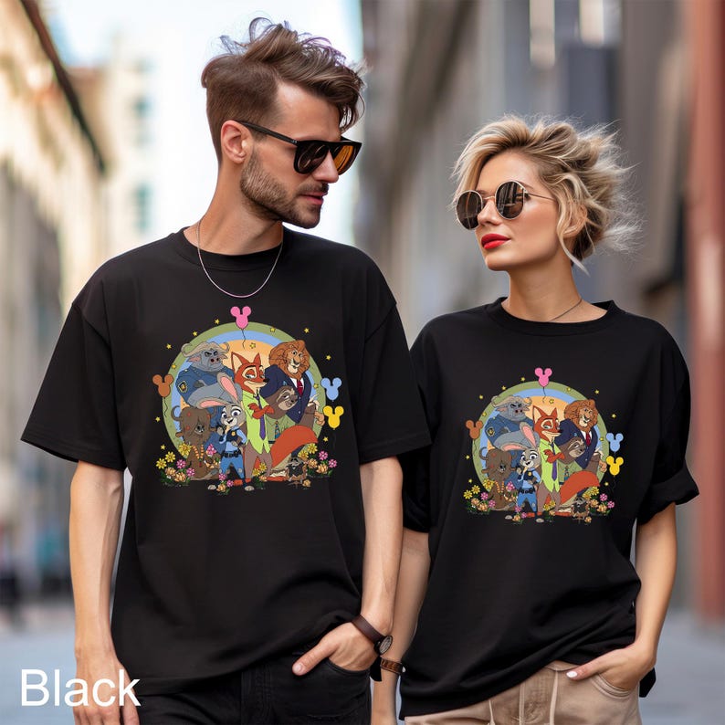 Pode incluir: T-shirts pretas com um gr&aacute;fico colorido de personagens de Zootopia. O design inclui Judy Hopps, Nick Wilde e outros personagens em um design circular com flores e &iacute;cones do Mickey Mouse. A palavra "Black" est&aacute; impressa na parte inferior.