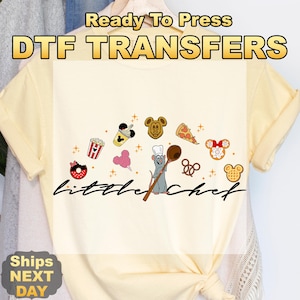 May include: Cream-colored t-shirt with a "little chef" design. The design includes a cartoon rat holding a wooden spoon, surrounded by food icons like popcorn, pizza, and Mickey Mouse-shaped treats. The text "Ready To Press DTF TRANSFERS" is at the top.
