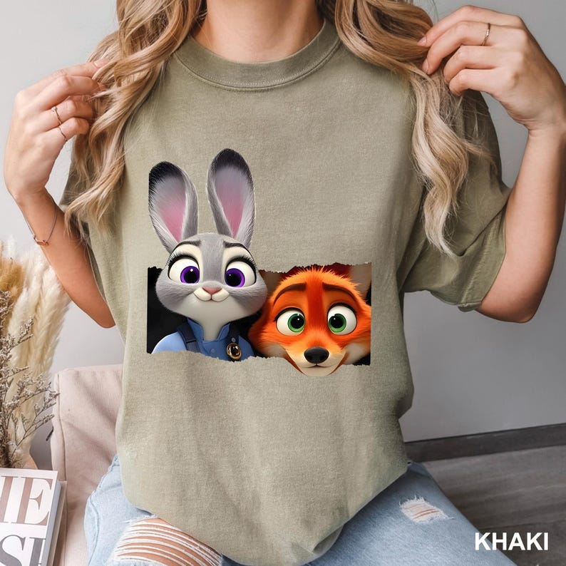 May include: Khaki-colored t-shirt featuring a graphic of Judy Hopps and Nick Wilde from Zootopia. The cartoon characters are in a rectangular frame. The word "KHAKI" is printed in white at the bottom right corner of the shirt.