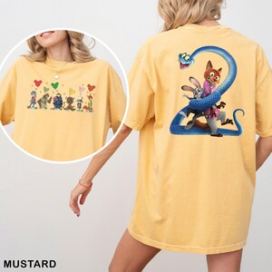 K&ouml;nnte beinhalten: Senffarbenes T-Shirt mit Cartoon-Grafiken auf Vorder- und R&uuml;ckseite. Die Vorderseite zeigt Charaktere mit herzf&ouml;rmigen Luftballons. Die R&uuml;ckseite zeigt einen Fuchs und ein Kaninchen mit einer blauen Schlange.