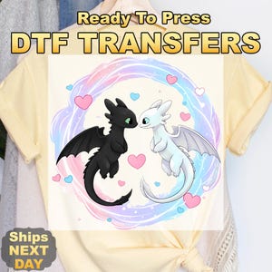 May include: A light yellow t-shirt featuring a DTF transfer design. The design includes two cartoon dragons, one black and one white, facing each other with hearts around them. The text "Ready To Press DTF TRANSFERS" is at the top.