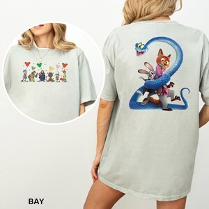 K&ouml;nnte beinhalten: Hellgr&uuml;nes T-Shirt mit Cartoon-Designs auf Vorder- und R&uuml;ckseite. Das Frontdesign zeigt Cartoon-Figuren mit herzf&ouml;rmigen Luftballons. Das R&uuml;ckendesign zeigt eine blaue Schlange mit Cartoon-Figuren. Das Wort "BAY" ist unten aufgedruckt.