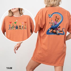K&ouml;nnte beinhalten: Orangefarbenes Kurzarm-T-Shirt mit einer Grafik auf der Vorderseite von Zeichentrickfiguren, die herzf&ouml;rmige Ballons halten. Auf der R&uuml;ckseite befindet sich eine Grafik eines Fuchses und eines Kaninchens mit einer blauen Schlange. Das Wort "YAM" ist unten aufgedruckt.