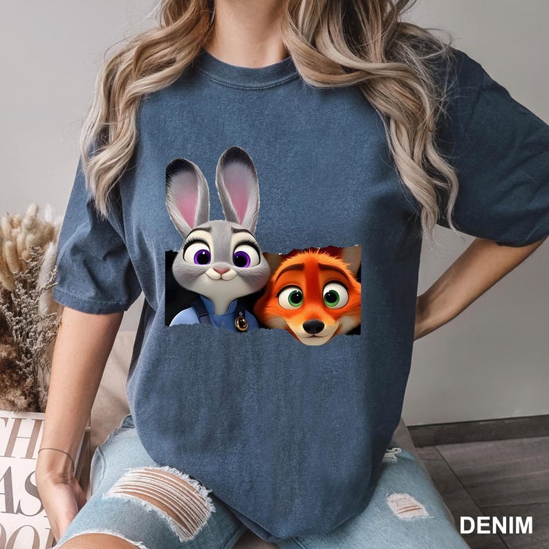 May include: Denim blue t-shirt featuring a graphic of Judy Hopps and Nick Wilde from Zootopia. The cartoon characters are in a rectangular design. The word "DENIM" is printed at the bottom right corner.