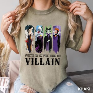 Puede incluir: Camiseta caqui con una ilustraci&oacute;n de cuatro villanos de dibujos animados y el texto "Sweetie I'm OK with being the Villain". La camiseta tiene cuello redondo y mangas cortas.
