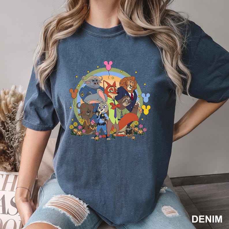 Pode incluir: Camiseta azul denim com um gr&aacute;fico colorido de personagens do filme de anima&ccedil;&atilde;o Zootopia, incluindo Judy Hopps e Nick Wilde, em um c&iacute;rculo floral. A palavra "DENIM" est&aacute; impressa na parte inferior.