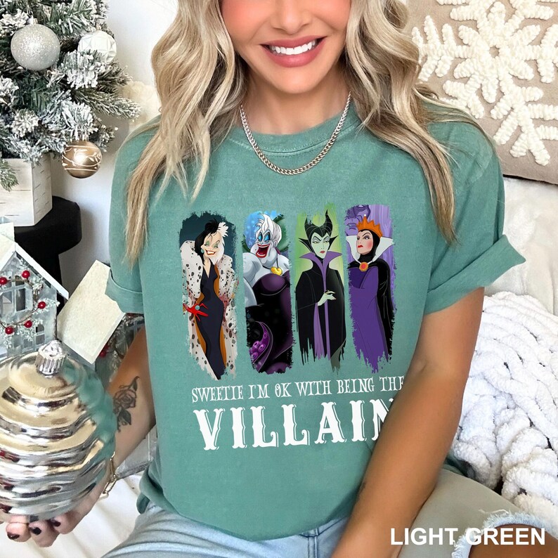 Puede incluir: Camiseta verde claro con un dise&ntilde;o de cuatro villanas de dibujos animados: Cruella de Vil, &Uacute;rsula, Mal&eacute;fica y la Reina Malvada. El texto "Sweetie I'm OK with being the Villain" est&aacute; impreso debajo.