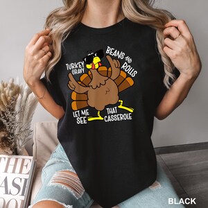 May include: Black t-shirt featuring a cartoon turkey wearing sunglasses and gesturing with its wings. The text on the shirt reads "Turkey Gravy, Beans and Rolls, Let Me See That Casserole."