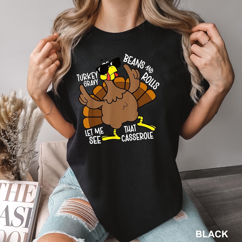 May include: Black t-shirt featuring a cartoon turkey wearing sunglasses and gesturing with its wings. The text on the shirt reads "Turkey Gravy, Beans and Rolls, Let Me See That Casserole."