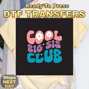 May include: A light yellow t-shirt featuring a black square graphic with the text COOL BIG SIS CLUB in wavy, retro-style lettering. The word COOL is pink with white flower accents, BIG SIS is purple, and CLUB is blue, all with small yellow stars.