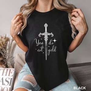 Peut inclure: T-shirt noir avec un motif d'&eacute;p&eacute;e blanche orn&eacute;e et le texte "You do not yield". Le motif comprend des &eacute;toiles et des fioritures d&eacute;coratives. Le t-shirt est &agrave; col rond.
