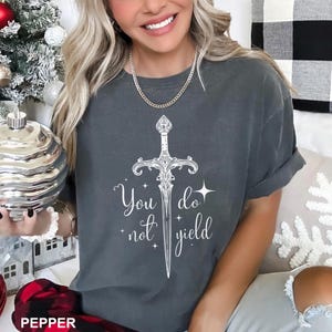 Peut inclure: T-shirt gris fonc&eacute; avec une &eacute;p&eacute;e blanche et le texte "You do not yield". Le design pr&eacute;sente une illustration d&eacute;taill&eacute;e d'&eacute;p&eacute;e avec des &eacute;l&eacute;ments d&eacute;coratifs. Le t-shirt est de style d&eacute;contract&eacute;.
