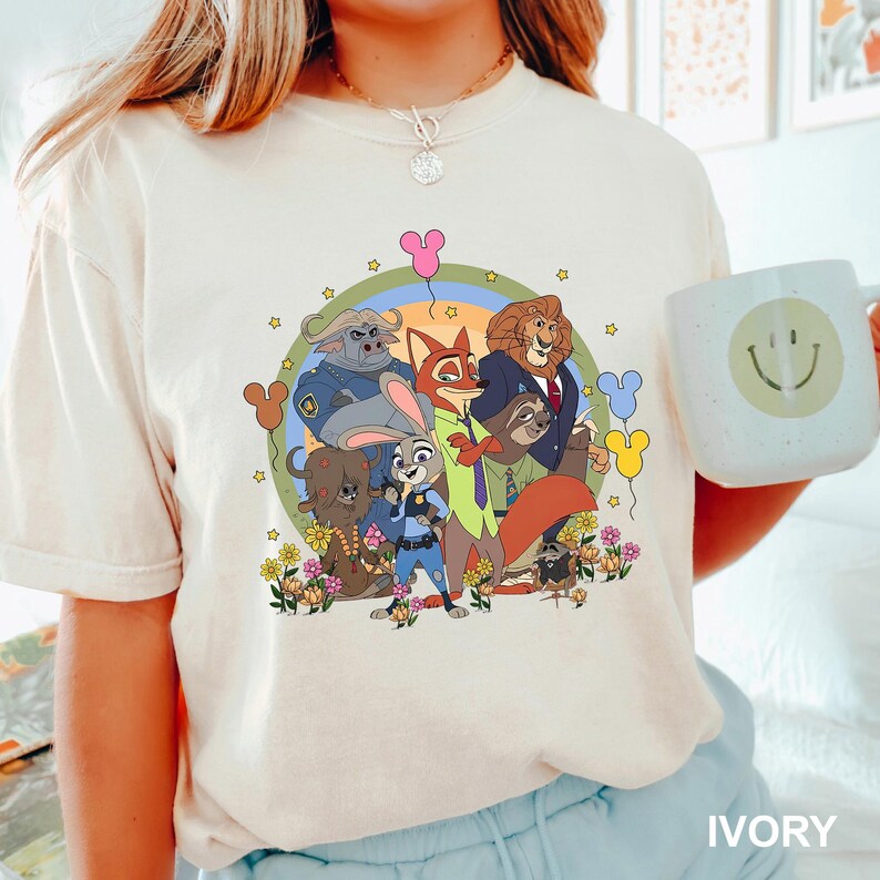 Pode incluir: Camiseta cor marfim com um gr&aacute;fico de desenho animado colorido de personagens de animais. O design inclui uma raposa, um coelho, um le&atilde;o e outros animais, rodeados por flores e bal&otilde;es. Uma caneca branca com um rosto sorridente &eacute; segurada na imagem.