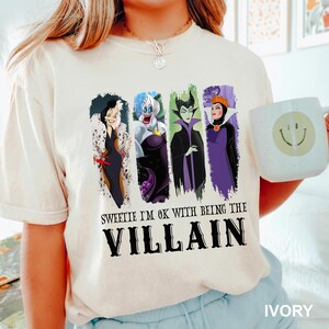 Puede incluir: Camiseta color marfil con cuatro personajes de villanos de dibujos animados. La camiseta tiene el texto "Sweetie I'm ok with being the Villain" en negro. Tambi&eacute;n se ve una taza blanca con una carita sonriente.