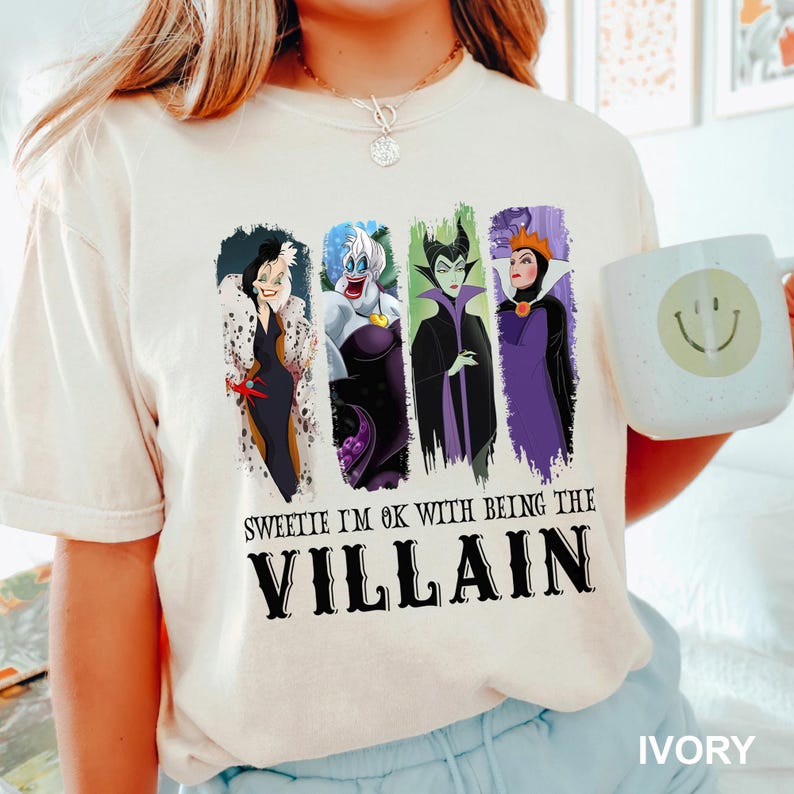 Puede incluir: Camiseta color marfil con cuatro personajes de villanos de dibujos animados. La camiseta tiene el texto "Sweetie I'm ok with being the Villain" en negro. Tambi&eacute;n se ve una taza blanca con una carita sonriente.