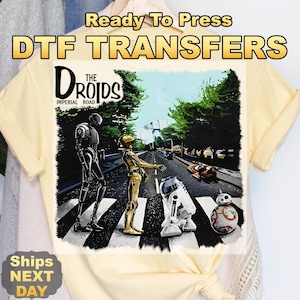 May include: Yellow t-shirt featuring a DTF transfer with a Star Wars theme. The graphic depicts droids crossing a street, with the text "The Droids Imperial Road" above the image. The words "Ready To Press" are at the top.