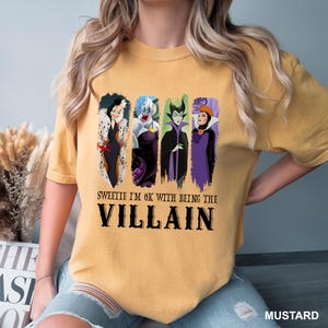 Puede incluir: Camiseta de color mostaza con cuatro personajes de villanos de dibujos animados. La camiseta tiene el texto negro "Sweetie I'm OK with being the Villain". La camiseta es de estilo casual.