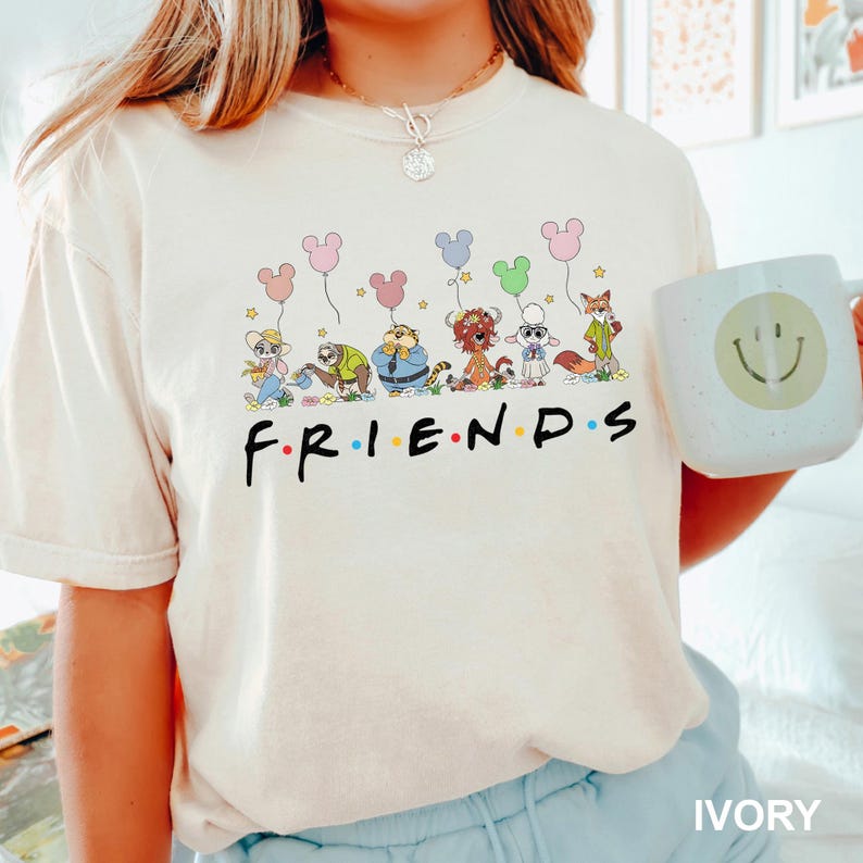 May include: Ivory-colored t-shirt featuring the word "FRIENDS" in a colorful font, with cartoon animal characters holding Mickey Mouse-shaped balloons. A white mug with a smiley face is visible. The shirt is a casual wear item.