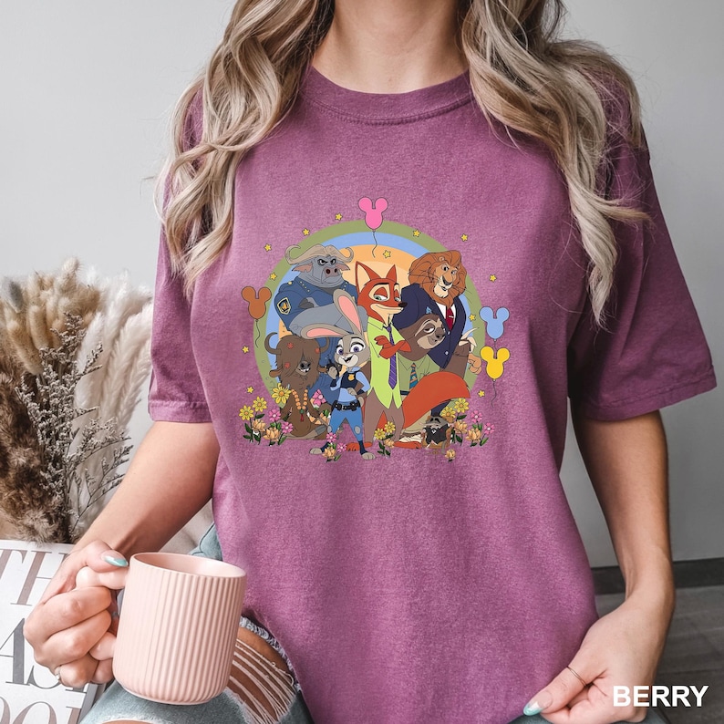 Pode incluir: Camiseta cor de baga com um gr&aacute;fico de desenho animado de personagens de Zootopia. O design inclui Judy Hopps, Nick Wilde e outros personagens, rodeados por flores e um arco-&iacute;ris. A camiseta tem gola redonda e mangas curtas.