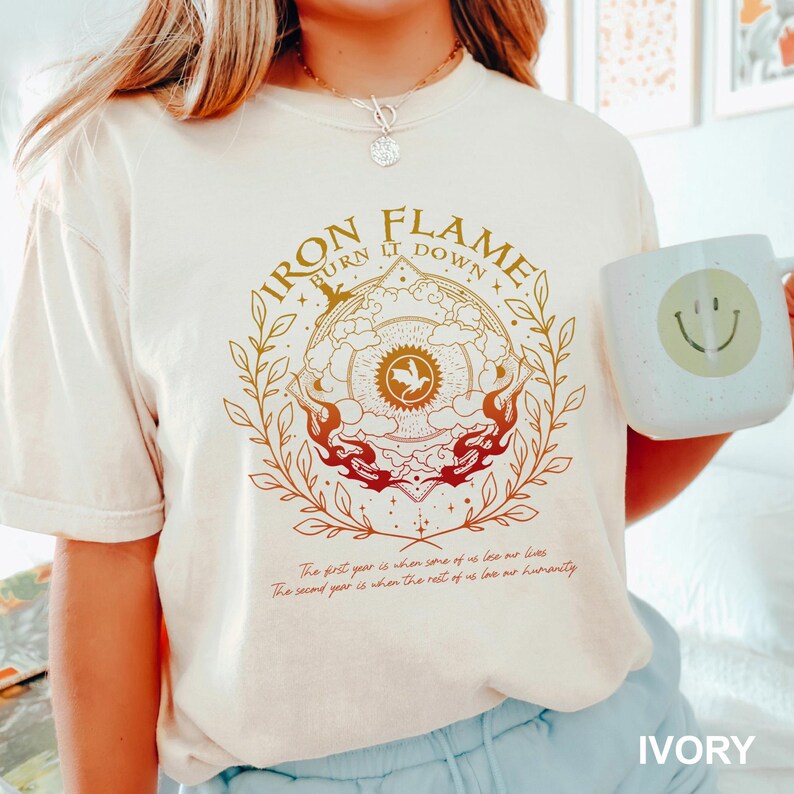 以下が含まれることがあります： アイボリー色のTシャツ。Iron Flame Burn It Downという文字と、炎、雲、月桂樹の冠に囲まれた中央のドラゴンのアイコンが描かれたグラフィックが特徴です。グラフィックの下にはThe first year is when some of us lose our livesというテキストが記されています。