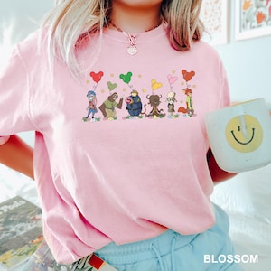 May include: A light pink crew neck sweatshirt with a cartoon graphic of animal characters holding heart-shaped balloons. The word "BLOSSOM" is printed in white at the bottom. A white mug with a smiley face is held in the hand.