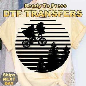 May include: A DTF transfer design featuring a black silhouette of a person riding a bicycle with a passenger against a striped background, with pine trees below. The text "Ready To Press DTF TRANSFERS" is at the top.