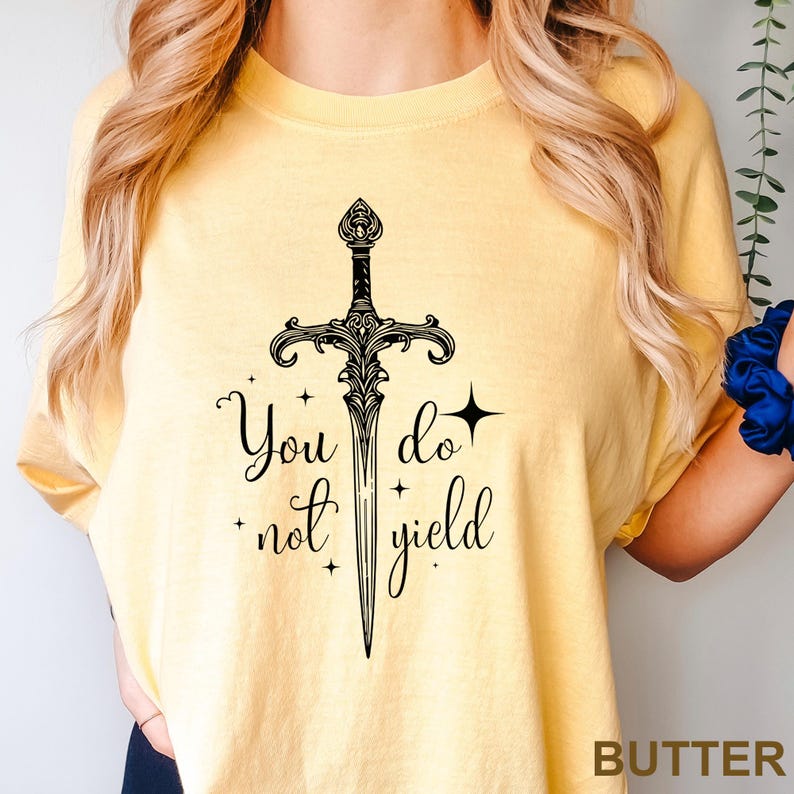 Peut inclure: T-shirt jaune clair avec une illustration d'&eacute;p&eacute;e noire et le texte "You do not yield". Le motif comprend des &eacute;toiles et des &eacute;l&eacute;ments d&eacute;coratifs. V&ecirc;tement d&eacute;contract&eacute;.