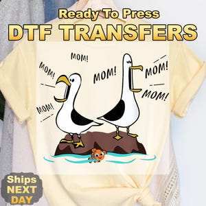 May include: DTF transfer featuring two cartoon seagulls with the word "MOM!" in speech bubbles. The seagulls are standing on a rock above blue water, with a small orange fish. The text "Ready To Press DTF TRANSFERS" is at the top.