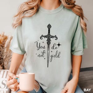 Peut inclure: T-shirt vert clair avec un motif d'&eacute;p&eacute;e noire orn&eacute;e et les mots "You do not yield". Le motif comprend des &eacute;toiles et des ornements d&eacute;coratifs. Le t-shirt est de couleur unie et a des manches courtes.