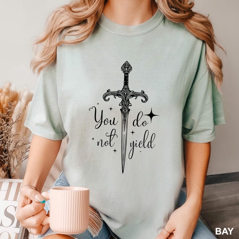 Peut inclure: T-shirt vert clair avec un motif d'&eacute;p&eacute;e noire orn&eacute;e et les mots "You do not yield". Le motif comprend des &eacute;toiles et des ornements d&eacute;coratifs. Le t-shirt est de couleur unie et a des manches courtes.