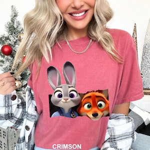 May include: Crimson-colored t-shirt featuring a graphic of Judy Hopps and Nick Wilde from Zootopia. The characters are depicted peeking out from a torn rectangle. The word "CRIMSON" is printed at the bottom of the shirt.